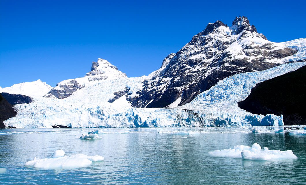 Snow-capped mountains and a glacier rise above a cold, blue lake scattered with floating ice chunks, offering breathtaking views from the nearby Singular Patagonia Hotel under a clear, bright blue sky.
