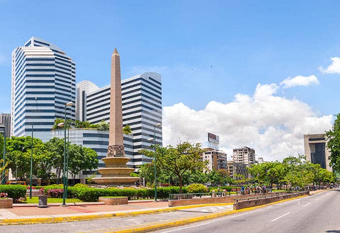 A cityscape with a tall stone obelisk in a landscaped plaza, modern striped glass buildings in the background, and a wide street lined with trees under a bright blue sky—perfect for exploring top things to do in Venezuela.