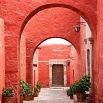 A narrow stone pathway with red walls and arches, lined with potted green plants, invites you to a wooden door in the background—the perfect trip setting for a unique travel experience.