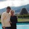 A man and woman stand together by a pool, smiling at the camera. The hills and a great cone-shaped structure rise in the background under a cloudy sky, making everything look picturesque.