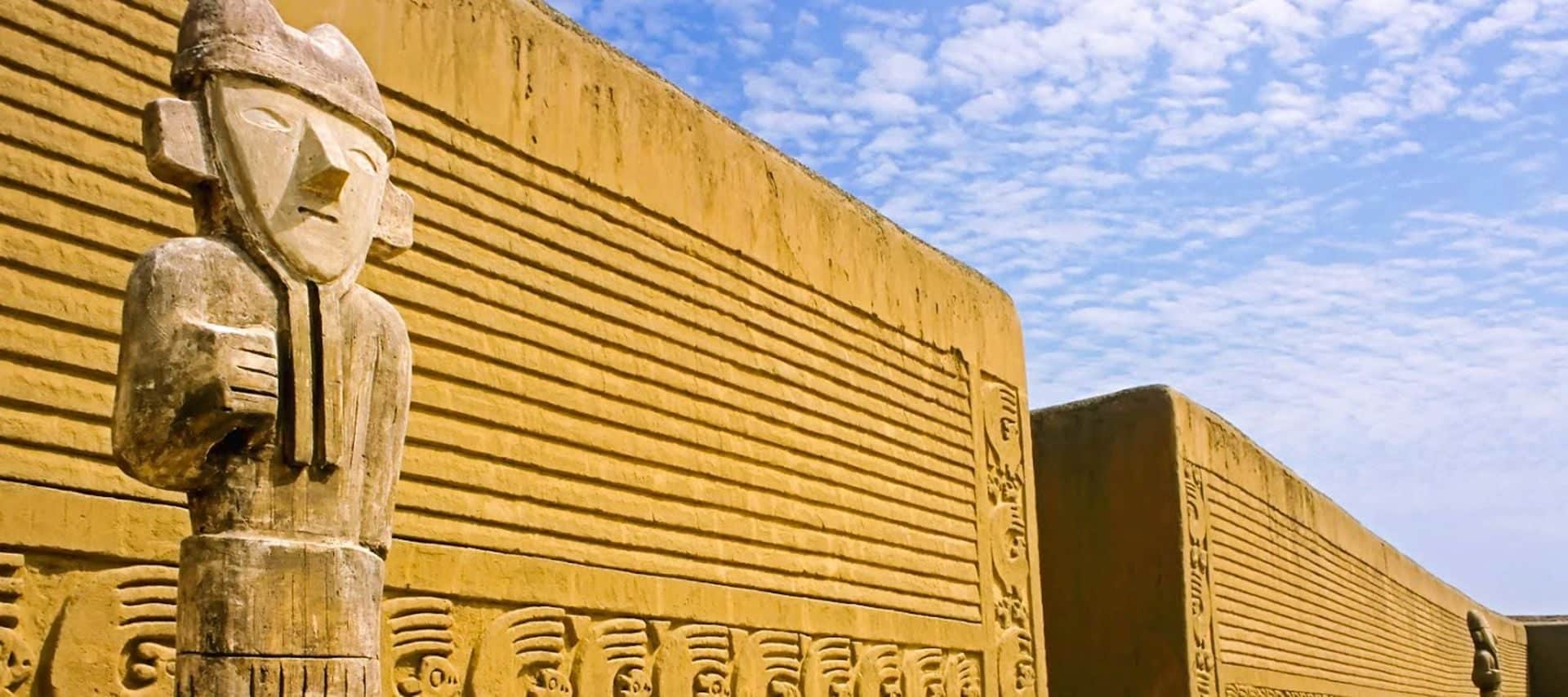 A carved stone statue stands in front of tall, yellow adobe walls adorned with geometric patterns under a partly cloudy blue sky at Chan Chan, Peru, capturing the spirit of ancient ruins often explored on archaeology tours.