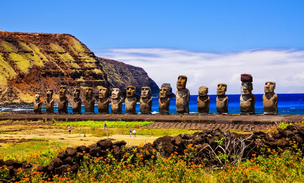 A row of large Moai statues stands on grassy terrain by the ocean on Rapa Nui, backed by cliffs and blue sky. People walk near the statues as orange wildflowers bloom in the foreground of Easter Island.