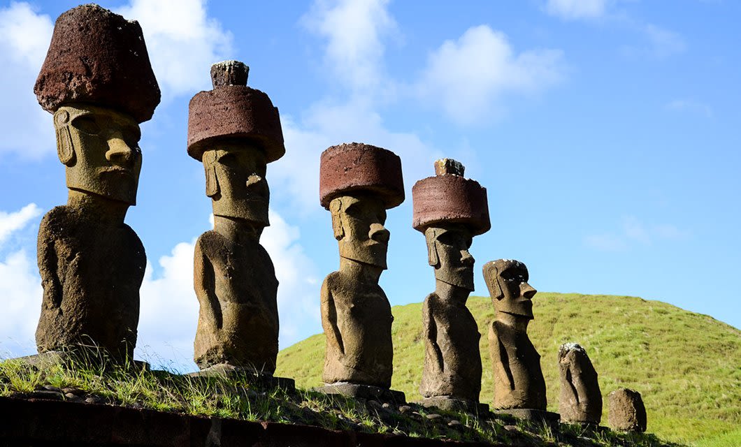 Six large stone Moai statues with red stone hats stand in a row on a grassy platform on Easter Island, beneath a blue sky—just steps from the eco-friendly comfort of Hangaroa Eco Village.
