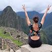 A person with arms raised sits on a ledge overlooking the ancient ruins of Machu Picchu in Peru, surrounded by the lush green mountains and misty skies that define this iconic Peruvian landscape.