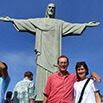 A man and woman smiling in front of the Christ the Redeemer statue in Rio de Janeiro, capturing memories of their fantastic trip with other tourists around and a clear blue sky in the background.