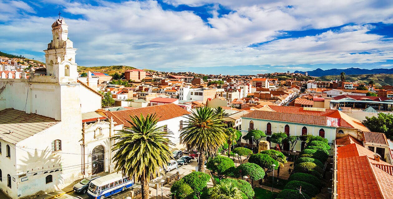 A vibrant cityscape with colonial-style buildings, a white church, palm trees, and a small park—an ideal setting for a Bolivia romantic getaway—unfolds under a bright blue sky dotted with scattered clouds.