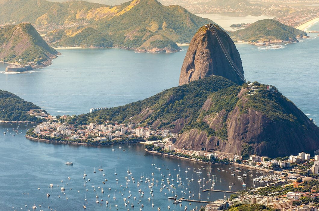 Aerial view of Sugarloaf Mountain in Rio de Janeiro, one of the top Brazil attractions, with boats in the harbor, lush green hills, and city buildings along the coastline under a bright, clear sky.