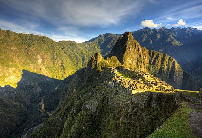 Aerial view of Machu Picchu, the ancient Incan city in Peru, surrounded by lush green mountains and sunlight. One of the top things to do here is explore its iconic ruins under a partly cloudy sky.