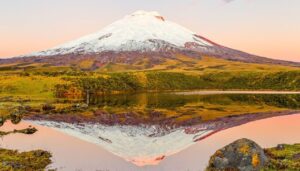 A snow-capped volcano rises above green hills, reflected clearly in a calm lake under a soft pink sky at sunset—one of the best places in Ecuador to experience breathtaking natural beauty.