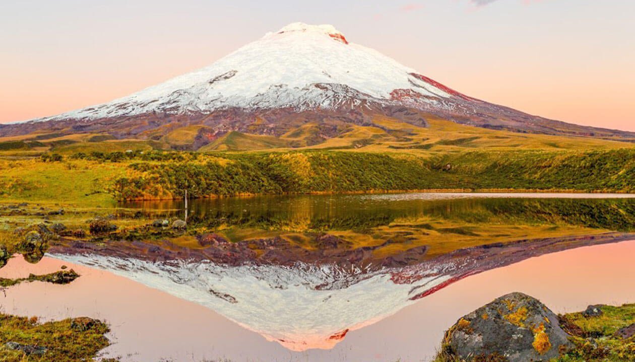 A snow-capped volcano rises above green hills, reflected clearly in a calm lake under a soft pink sky at sunset—one of the best places in Ecuador to experience breathtaking natural beauty.