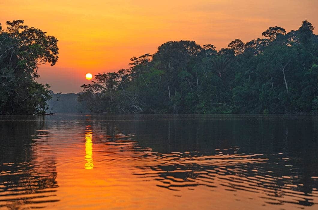 The sun sets over a calm river surrounded by dense green trees, casting an orange and yellow reflection on the water—a breathtaking scene that showcases the natural beauty of Brazil attractions.