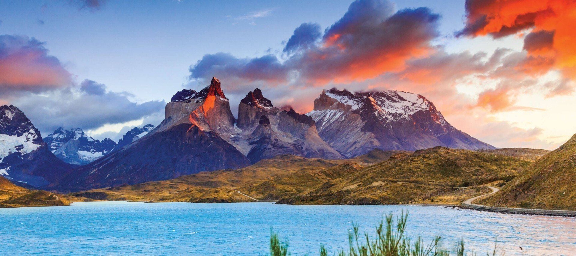 Snow-capped mountains rise dramatically behind a turquoise lake under a colorful sky at sunrise or sunset—this stunning scene captures the magic of South America travel, especially during the best time to visit for breathtaking views.