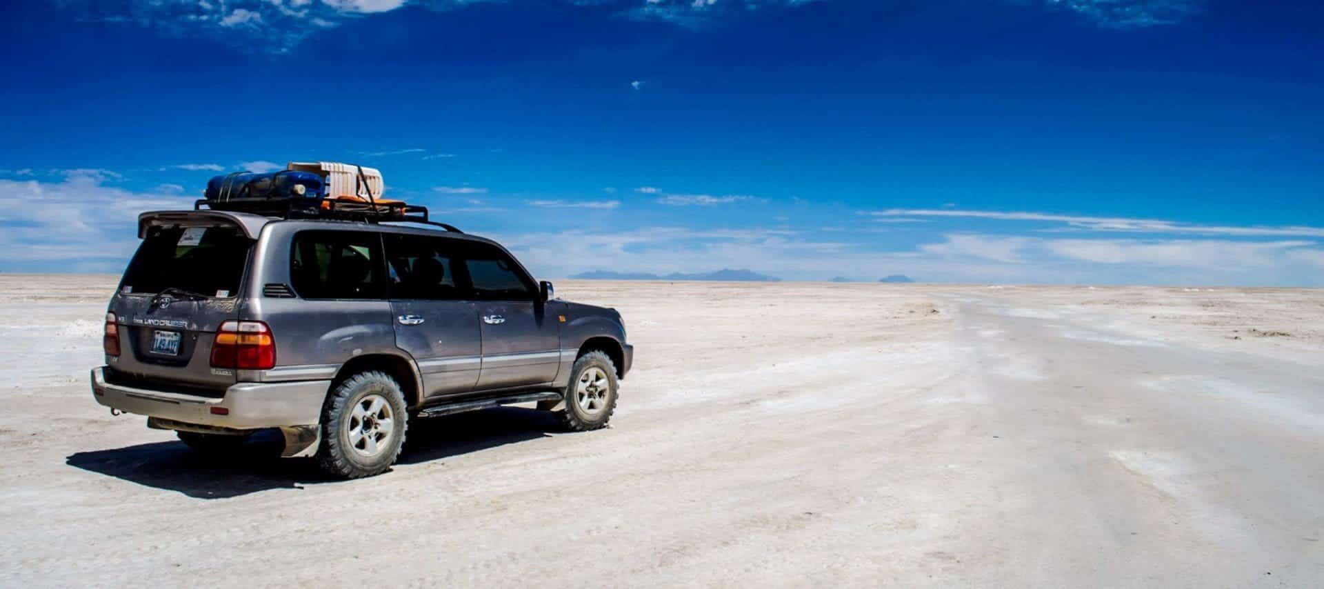 A silver SUV with luggage on the roof is parked on Bolivia’s vast, flat, white desert landscape under a bright blue sky with scattered clouds—perfect for unforgettable adventure tours across the distant, empty horizon.