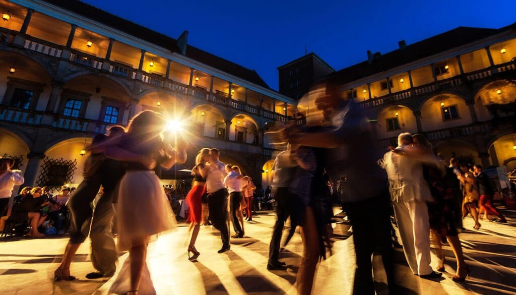 Couples dance together in a well-lit outdoor courtyard at dusk, surrounded by an illuminated historic building with arches and balconies—a lively, festive scene reminiscent of top festivals in Argentina.