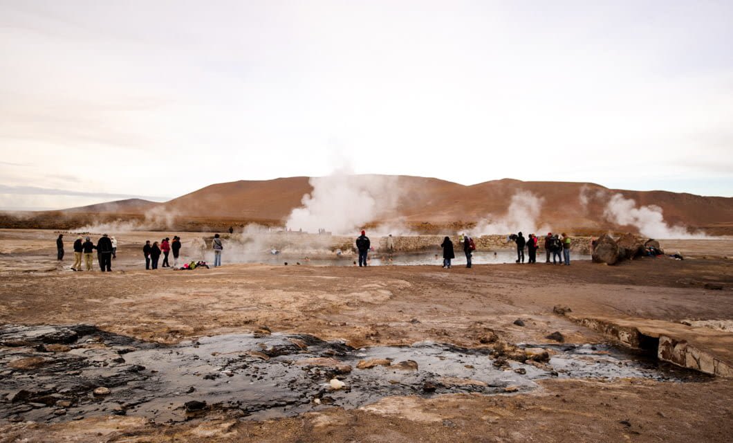 A group of people stand near steaming geysers on a rocky, barren landscape with distant hills under a cloudy sky, exploring the raw beauty near Casa Atacama.