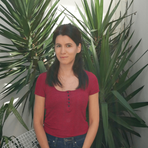 A woman with long dark hair, wearing a red short-sleeve shirt and jeans, stands in front of tall green plants indoors, smiling slightly at the camera.