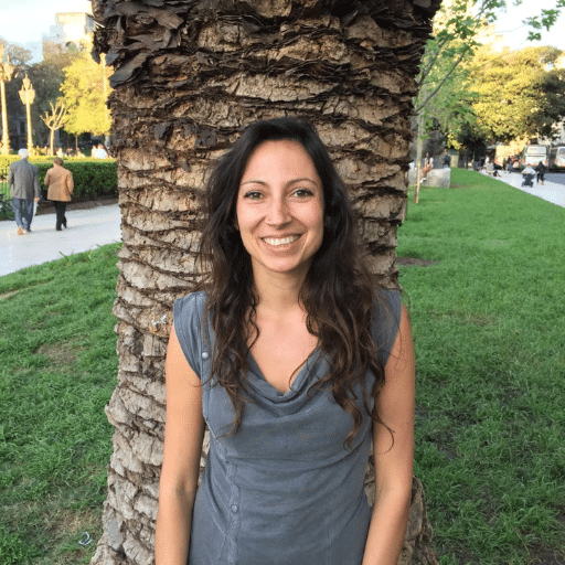 A woman with long brown hair, wearing a gray sleeveless top, smiles while standing in front of a palm tree in a park with green grass and people walking in the background.