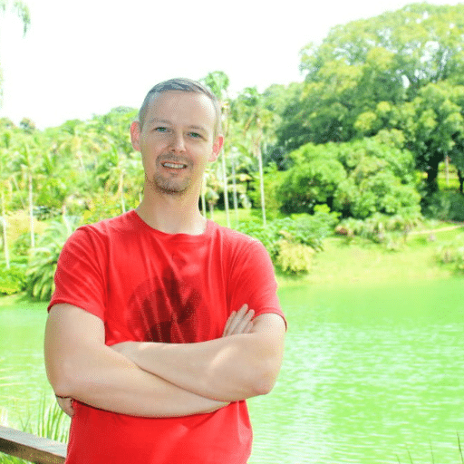 A man in a red t-shirt stands with arms crossed, smiling in front of a green lake and lush trees on a sunny day.