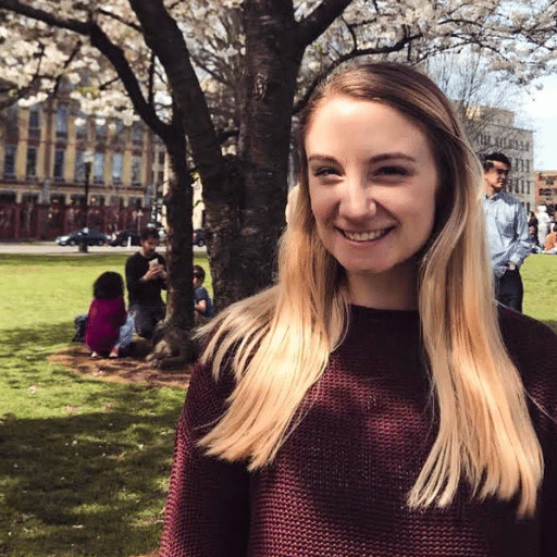 A young woman with long blonde hair and a maroon sweater smiles in a sunny park, standing in front of a tree with blooming white flowers. People sit and walk on the grass in the background.