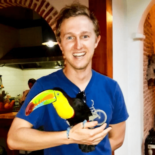 A smiling person in a blue t-shirt holds a colorful toucan inside a warmly lit room with arched doorways and brick walls.