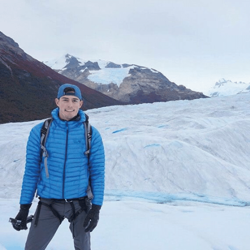 A man in a blue jacket and cap stands on a snowy, icy landscape with mountains in the background, smiling at the camera and holding a camera in his right hand.