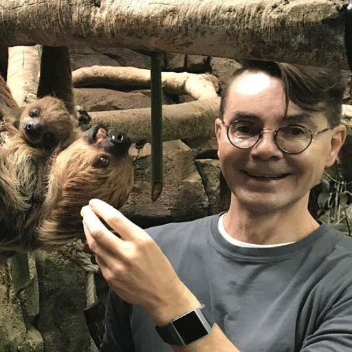 A smiling person wearing glasses poses next to a sloth and its baby hanging upside down from a tree branch in an indoor animal exhibit.