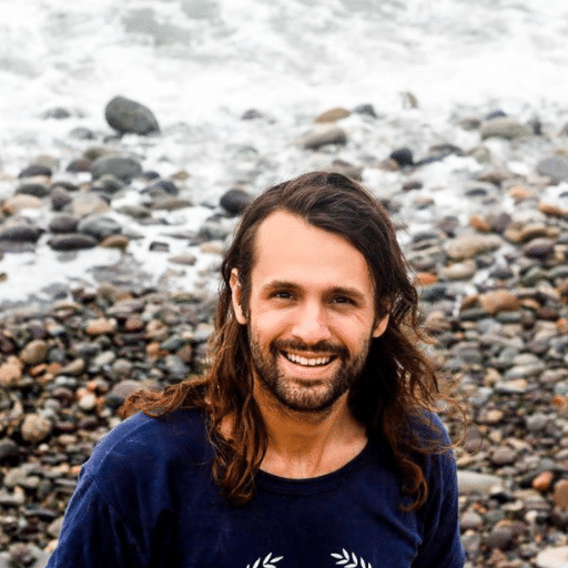 A man with long brown hair and a beard smiles at the camera while standing on a rocky beach, with waves and stones visible in the background.
