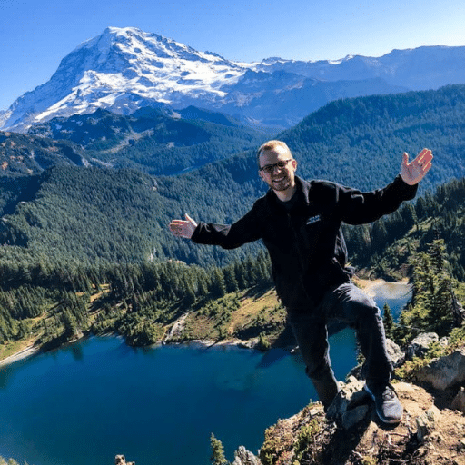 A smiling man with glasses poses on a rocky ledge overlooking a blue lake and dense forest, with a snow-capped mountain rising majestically in the background under a clear blue sky.