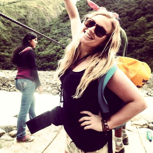 A smiling woman with long blonde hair, wearing sunglasses and a backpack, poses outdoors in front of a river and green hills. Another woman stands nearby, looking away. The atmosphere is adventurous and sunny.