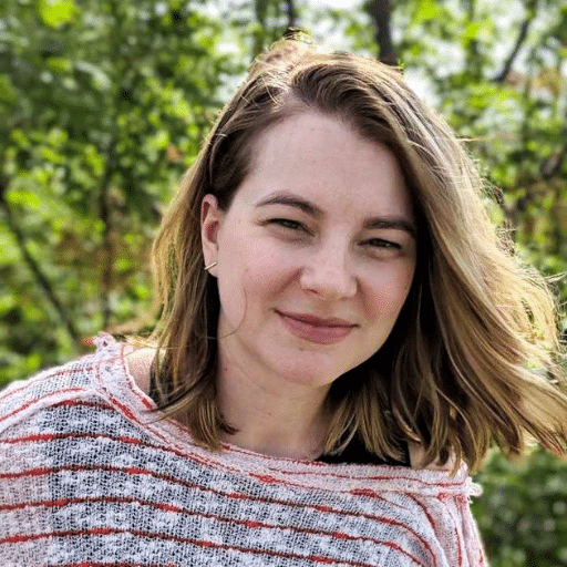 A woman with light brown hair and a striped sweater smiles softly outdoors, with green trees and sunlight in the background.
