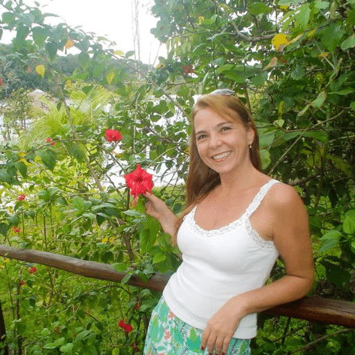 A woman with light brown hair, wearing a white tank top and patterned skirt, smiles while standing outdoors by green foliage and red flowers, holding a wooden railing.