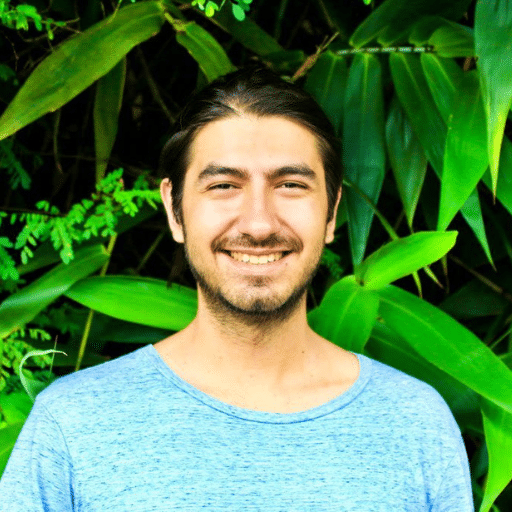 A young man with long dark hair tied back, wearing a light blue shirt, smiles in front of a lush green background of large tropical leaves.