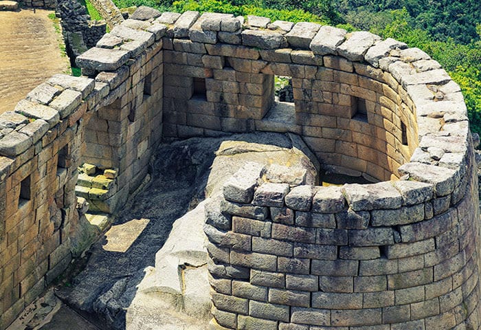 An ancient stone structure with curved walls and rectangular windows, set on a rocky foundation amidst greenery. The weathered stones are tightly fitted, showcasing historic Inca architecture at Machu Picchu—a must-see for travel enthusiasts.