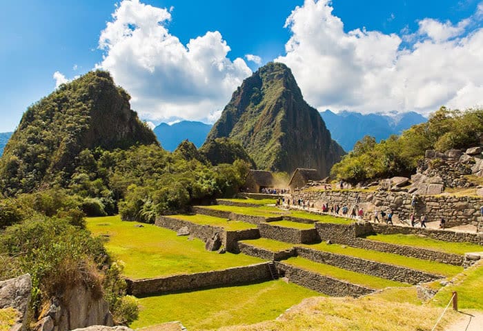 Ancient stone terraces and ruins of Machu Picchu sit atop a lush green mountain with dramatic peaks and a partly cloudy blue sky. Tourists explore the iconic site, discovering unforgettable things to do along its scenic paths.