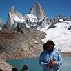 A person wearing a hat and blue shirt stands near a turquoise glacial lake, surrounded by snow-capped, jagged mountain peaks under a clear blue sky, capturing memorable moments in nature’s grandeur.