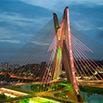 Cable-stayed bridge with two large towers lit by colorful lights, crossing a river in Argentina, with city buildings and streetlights visible in the background at dusk—a captivating view for any trip or travel adventure.