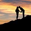 Two people stand on a hill at sunset during their trip, one leaning in to kiss the other on the forehead, creating a silhouette against the colorful sky.
