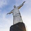 A low-angle view of the Christ the Redeemer statue in Rio de Janeiro, Brazil, with arms outstretched against a blue sky with light clouds, captured as part of a professional work showcasing iconic landmarks.