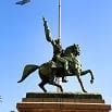 A wonderful bronze equestrian statue of a man raising one arm while riding a horse stands on a stone pedestal, set against a clear blue sky—an inspiring sight for any travel enthusiast.