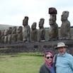 A man and woman stand in front of a row of large stone moai statues on Easter Island, enjoying the grassy landscape and cloudy sky during their fantastic trip.