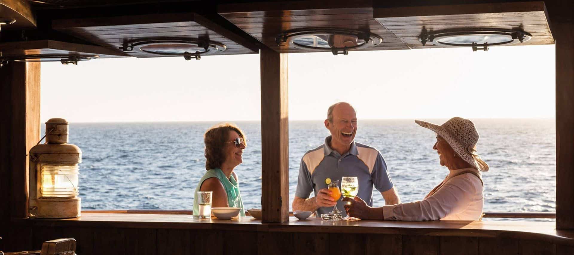 Three older adults smiling and enjoying drinks at an outdoor bar on a ship during an Ecuador Cruises voyage, with the ocean and bright sunlight in the background.