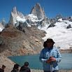 A person in a sunhat and blue jacket stands by a turquoise glacial lake, surrounded by snow-covered mountains and jagged peaks. On this clear day, others gather at the lake’s edge, creating memorable moments in this breathtaking setting.