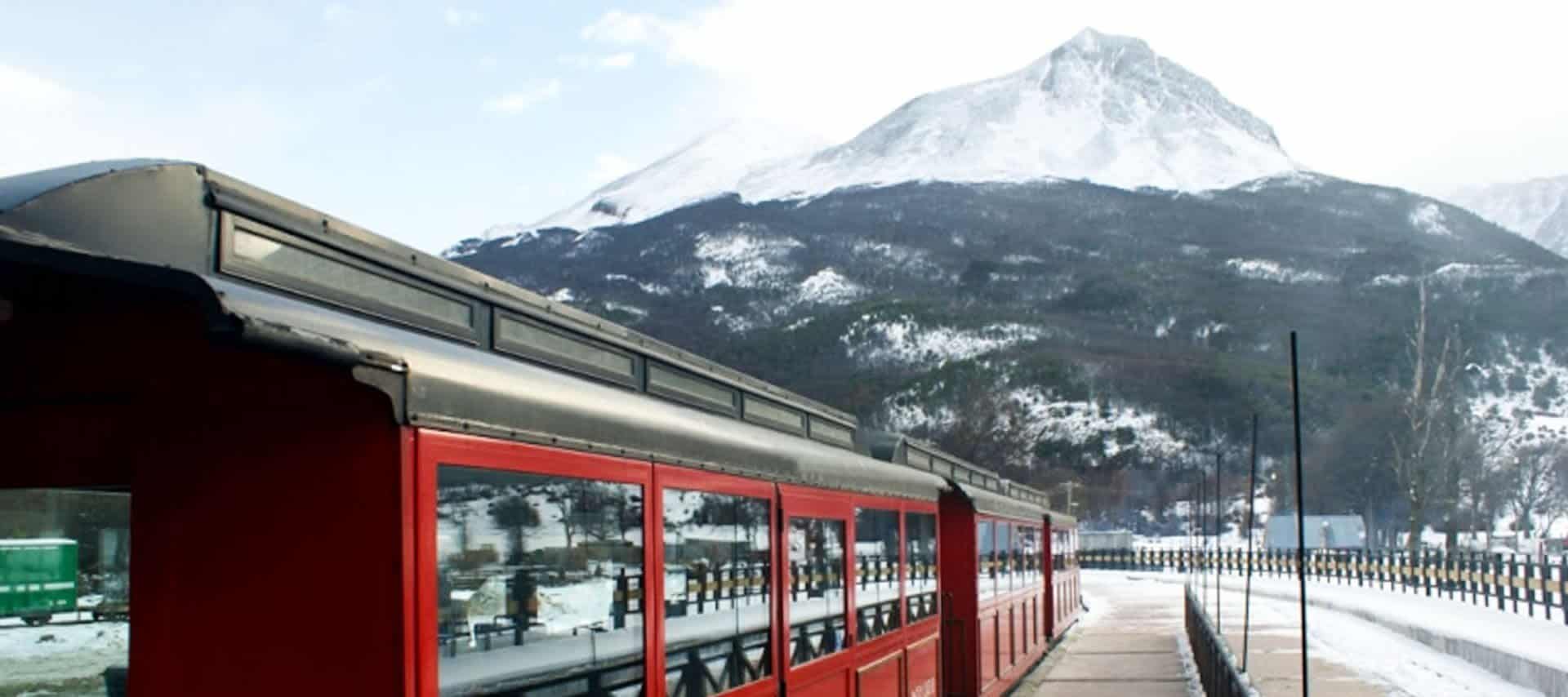 A red train stands at a snowy station platform with a snow-covered mountain and forested slopes in the background under a partly cloudy sky, capturing the spirit of Patagonia Train Tours.