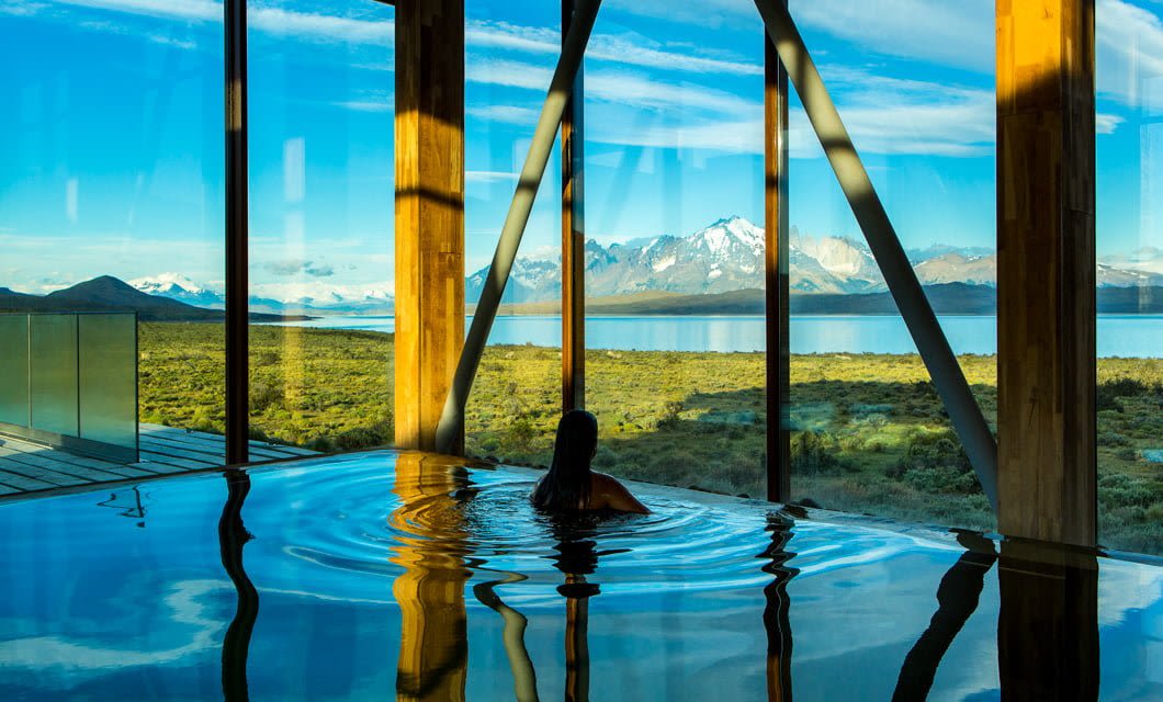 A person relaxes in an indoor pool at Tierra Patagonia, with large windows overlooking grassy plains, a tranquil lake, and snow-capped mountains beneath a blue sky.