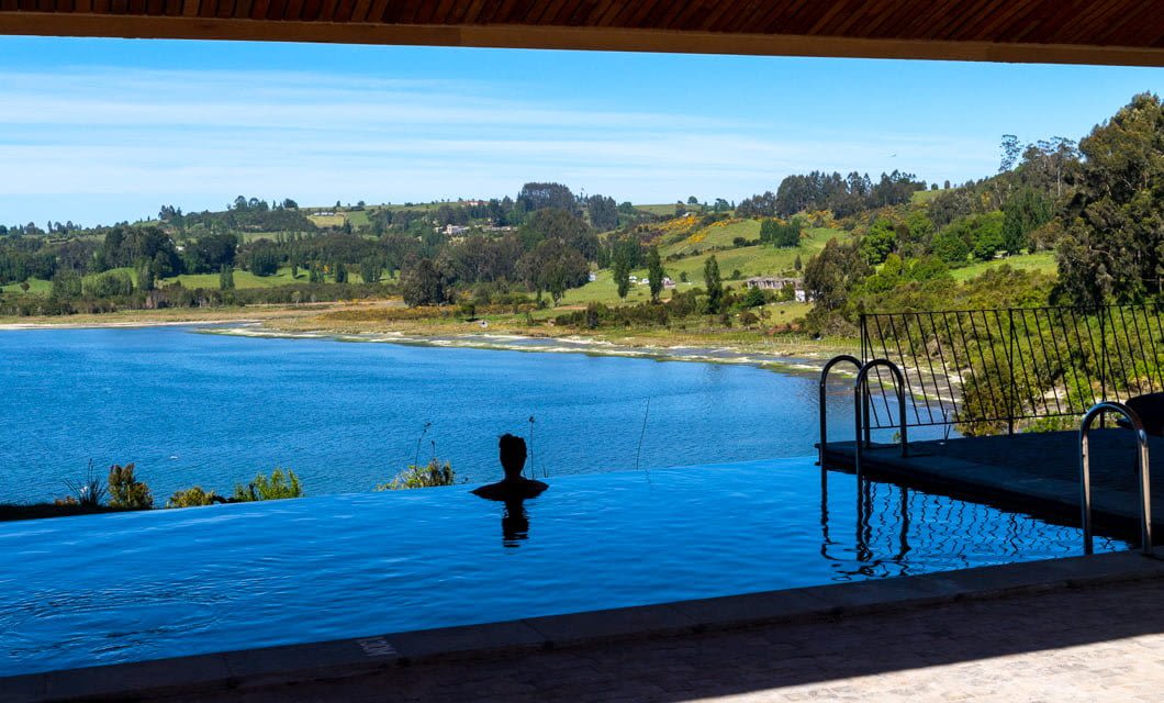 A person relaxes in an infinity pool at Refúgia Hotel, overlooking a blue lake with green hills, trees, and scattered houses in the background under a clear sky.