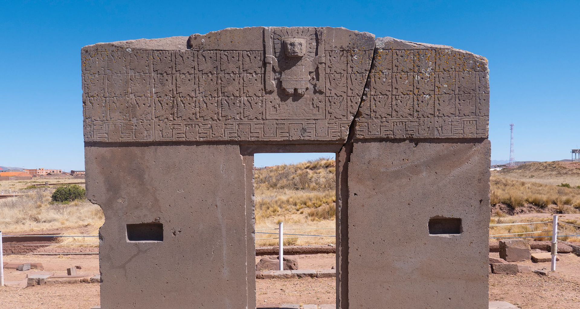 A large ancient stone gateway, known as the Gate of the Sun, featuring intricate carvings and a central deity figure at the top, stands in a dry, grassy Bolivian landscape under a clear blue sky—a UNESCO marvel awaiting discovery.