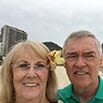 An older woman and man smile at the camera on a beach in Brazil. The woman has a yellow flower in her hair, with tall buildings and cloudy skies visible in the background.