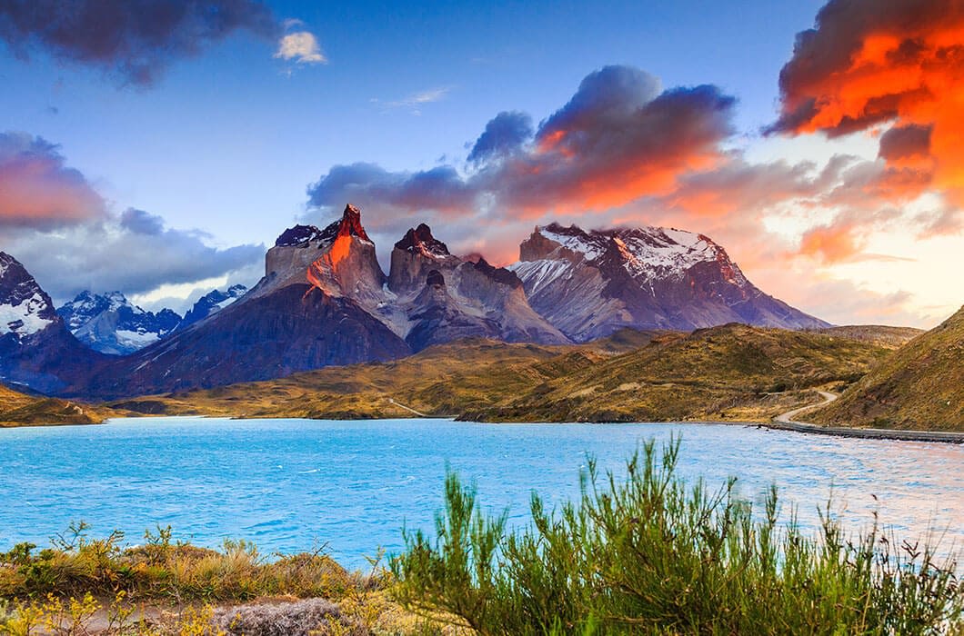 A bright blue lake sits before rugged, snow-capped mountains under a dramatic sunset sky. Bushes and grassy terrain frame the scene—an unforgettable view among top Chile attractions for those seeking memorable Chile travel experiences.