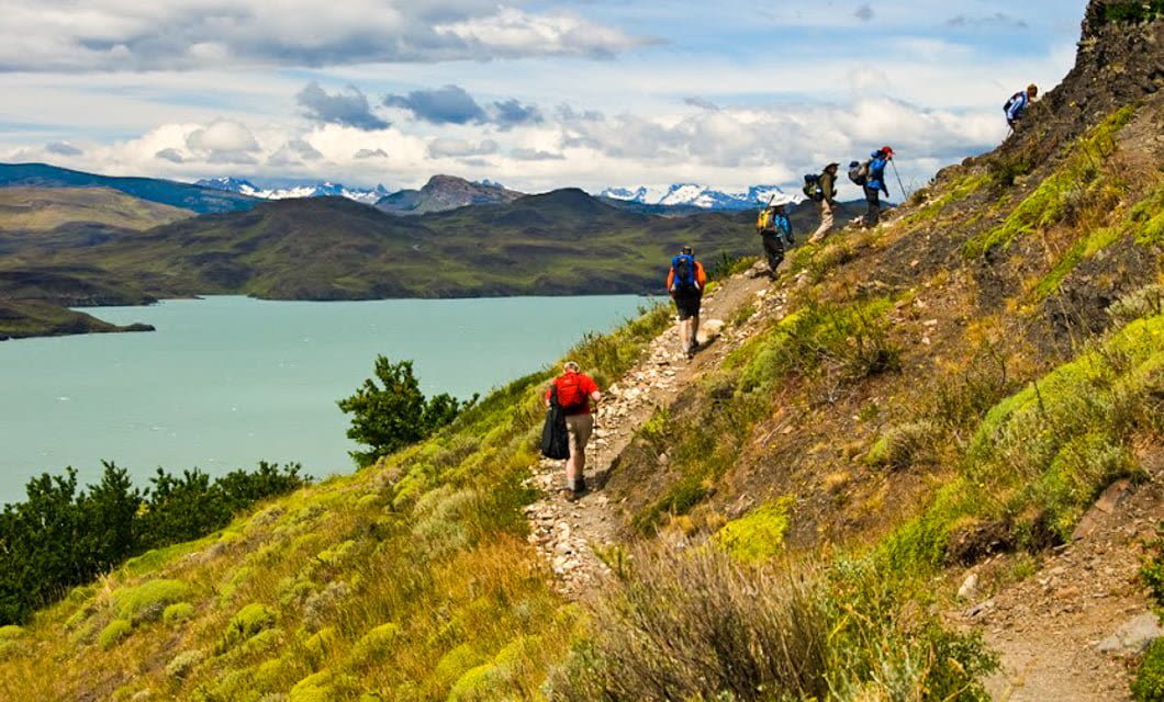 A group of hikers with backpacks walk along a narrow, rocky path on a hillside in Patagonia, overlooking a turquoise lake and distant snow-capped mountains under a partly cloudy sky—an iconic scene of adventure travel.
