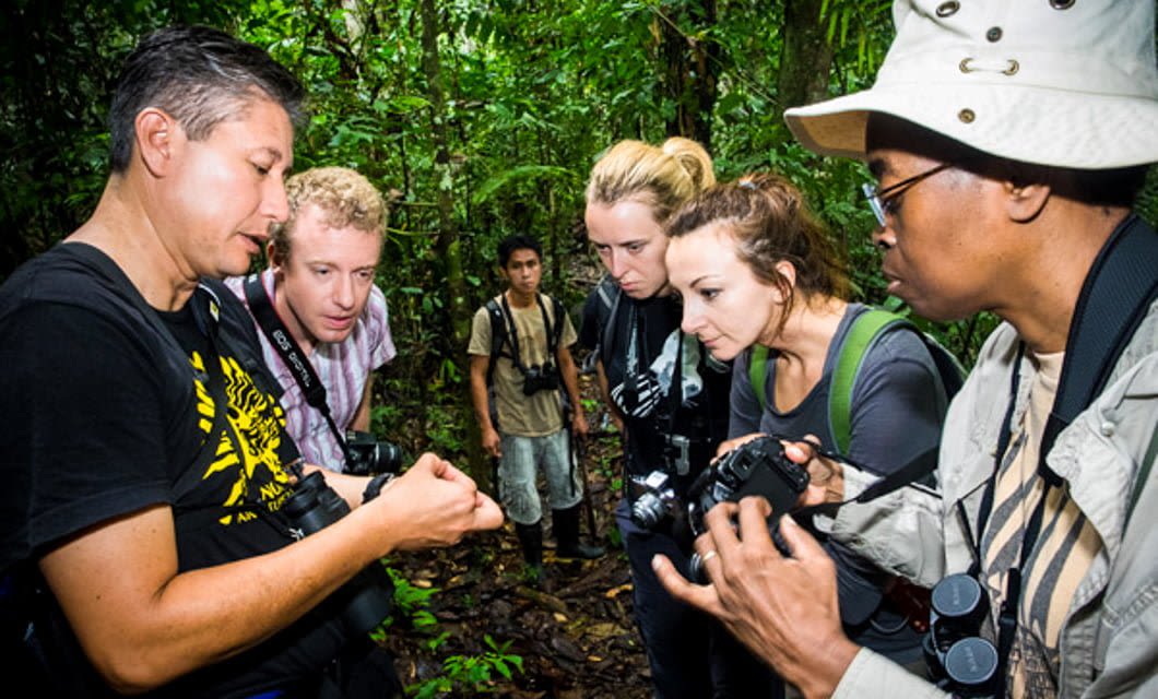 A group of people in the Amazon rainforest closely observe something in a man's hand, with cameras and binoculars ready, while another person stands in the background.
